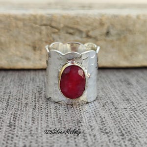 May include: A silver ring featuring a red gemstone set in a hammered band. The ring has a wavy edge and is a statement piece.