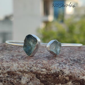 May include: A silver bangle bracelet with two light blue aquamarine stones. The stones are set in silver and are a raw, natural shape.