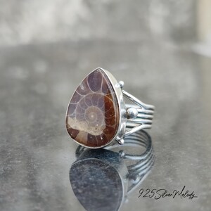 Peut inclure: Une bague en argent avec une pierre pr&eacute;cieuse ammonite en forme de larme. La bague a un design d&eacute;licat et complexe avec une bande en argent et un petit accent de perle en argent.