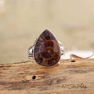 Peut inclure: Une bague en argent avec une pierre pr&eacute;cieuse en ammonite en forme de larme. L'ammonite est brune et pr&eacute;sente un motif en spirale. La bague est pos&eacute;e sur une surface en bois.