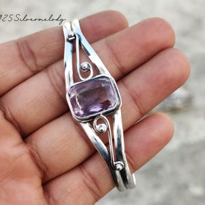 Peut inclure: Un bracelet jonc en argent avec une pierre précieuse améthyste sertie dans un motif décoratif. Le bracelet est ouvert et présente un design délicat et complexe.