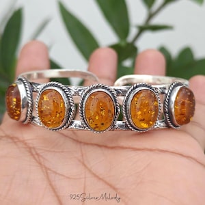 May include: A sterling silver cuff bracelet featuring five oval-shaped amber stones set in twisted rope-style bezels. The silver band has a textured, hammered finish. The text 925 Silver Melody is printed at the bottom of the image.