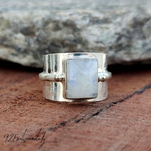 May include: A silver ring featuring a rectangular moonstone. The ring has a wide band with decorative elements and a polished finish. The ring is displayed on a brown surface.