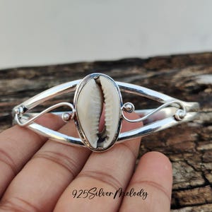 Peut inclure: Un bracelet en argent avec un coquillage de cauris blanc au centre. Le bracelet a un design délicat et orné.