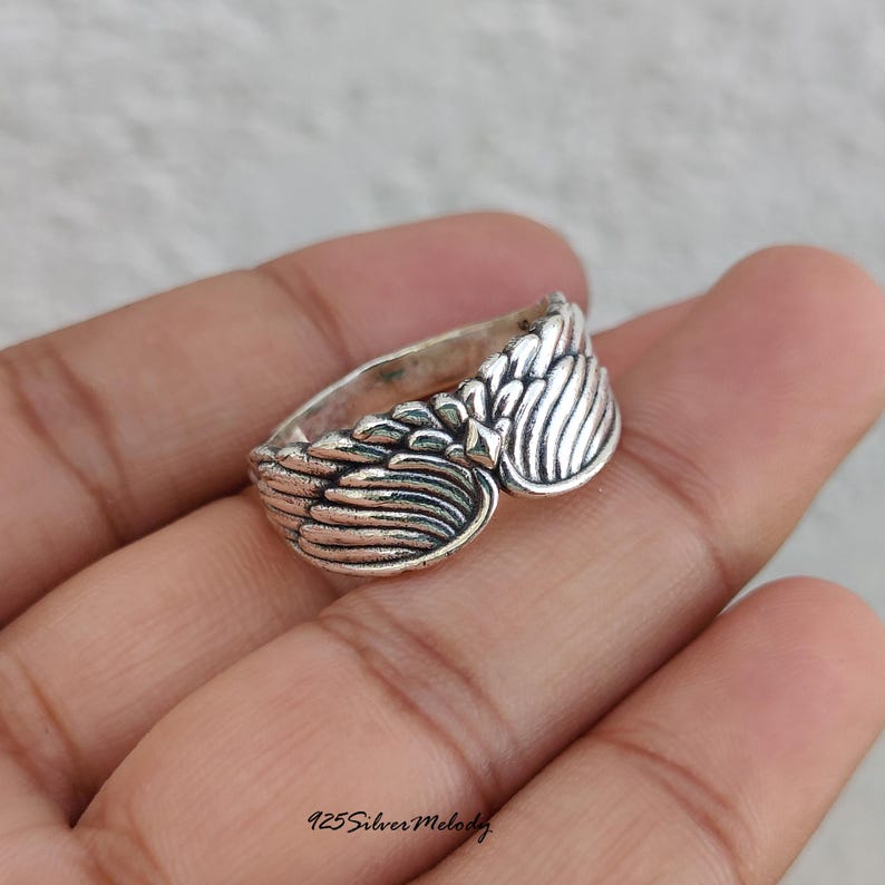 May include: A silver ring featuring a detailed wing design that wraps around the band. The metal has an oxidized finish to highlight the texture of the feathers. The text 925 Silver Melody is printed in the bottom corner of the image.