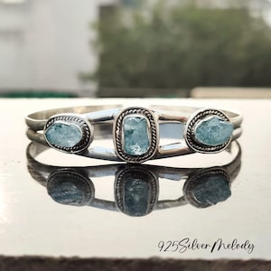 May include: A silver cuff bracelet featuring three light blue aquamarine stones. The stones are set in a silver band with a rope-like border. The bracelet is reflected on a shiny surface. The text "925 Silver Melody" is at the bottom.