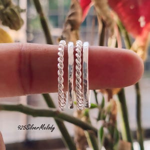 Puede incluir: Cuatro anillos de plata apilados en un dedo. Los anillos son todos finos y delicados, dos de ellos tienen un diseño de cuerda trenzada y dos de ellos tienen un acabado liso y martillado.
