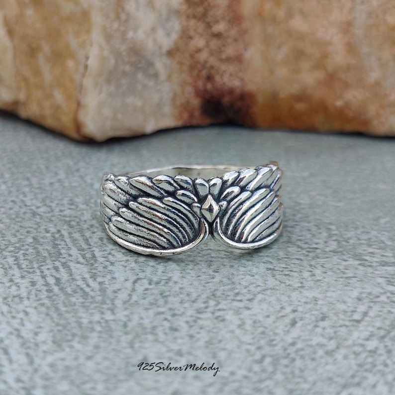 May include: A sterling silver ring featuring a detailed wing design that wraps around the band. The metal has an oxidized finish to highlight the textured feather patterns and a small diamond-shaped accent in the center. Text reads 925SilverMelody.