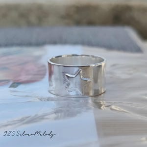 May include: A silver ring with a star design. The ring is wide and has a hammered texture. The text "925 Silver Melody" is visible in the bottom left corner of the image.