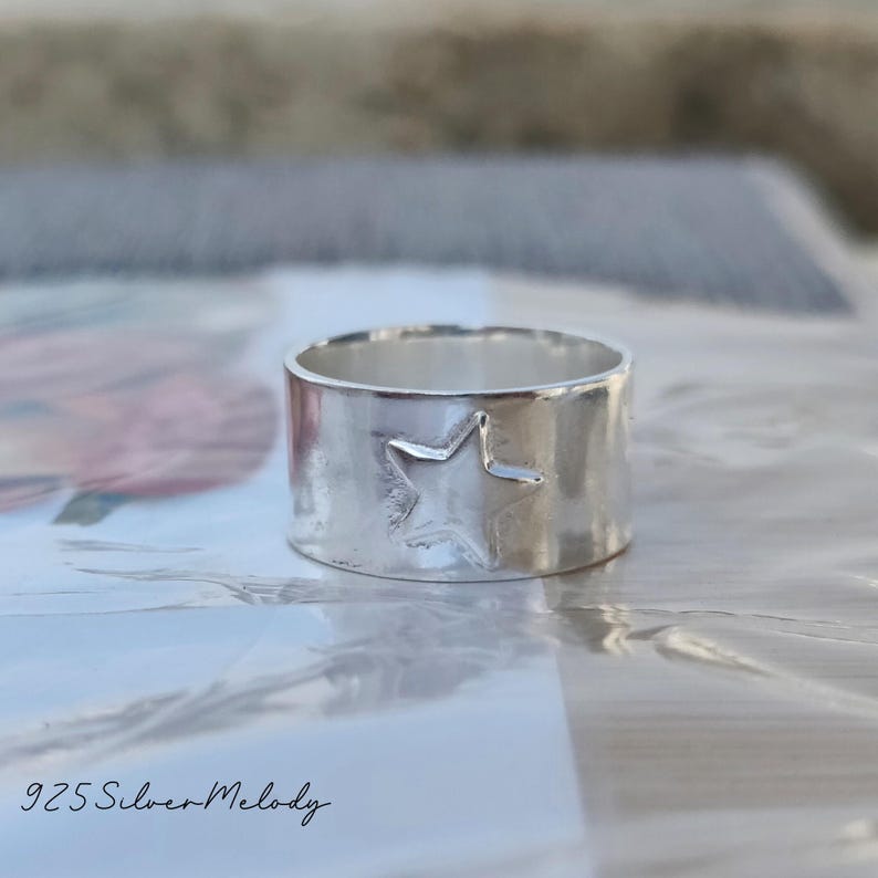May include: A silver ring with a star design. The ring is wide and has a hammered texture. The text "925 Silver Melody" is visible in the bottom left corner of the image.