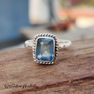 Peut inclure: Une bague en argent avec une pierre précieuse bleue sertie dans un bézel carré. Le bézel est orné d'un motif de corde autour du bord.