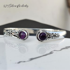 May include: A silver bangle bracelet with two amethyst gemstones. The bracelet features a delicate floral design.