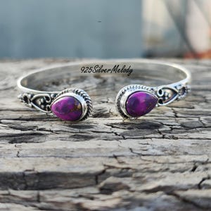 May include: A silver cuff bracelet with two purple and gold teardrop-shaped stones. The bracelet is open on both ends and has a delicate design.