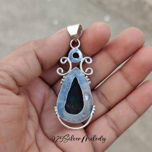 K&ouml;nnte beinhalten: Silberner Anh&auml;nger mit einem dunkelgr&uuml;nen Edelstein, der in ein filigranes Silberdesign eingefasst ist. Der Anh&auml;nger befindet sich auf einer Hand, und der Text "925 Silver Melody" ist am unteren Rand des Bildes sichtbar.