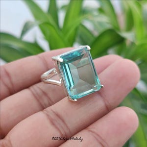 May include: A silver ring featuring a large, rectangular, faceted light blue gemstone held by four prongs. The band has a split design, and the text 925 Silver Melody is printed in black at the bottom of the image, indicating the metal composition.