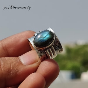 Peut inclure: Une bague en argent avec une grande pierre ovale de labradorite bleue sertie dans une large bande. La bande est décorée de motifs floraux complexes.