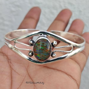 Può includere: Un bracciale a fascia in argento sterling con una pietra ovale iridescente al centro con riflessi verdi, rossi e arancioni. La fascia in argento lucido ha un design diviso con sfere metalliche decorative accanto alla pietra. Testo: 925SilverMelody.