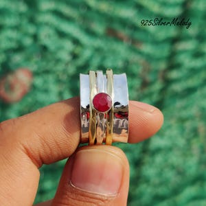 May include: A silver and gold spinning ring with a red gemstone. The ring is worn on a finger.