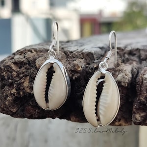 May include: A pair of silver earrings featuring a white cowrie shell design. The earrings are hanging from a wooden surface.