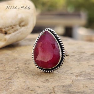 May include: A silver ring featuring a large, pear-shaped, faceted ruby gemstone. The ruby is a deep red color and is set in a silver band with a twisted rope design around the stone. The text "925 Silver Melody" is in the upper left corner.