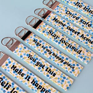 Pode incluir: Chaveiros com padrões florais e afirmações positivas. Cada chaveiro apresenta um fundo branco com desenhos florais azuis e amarelos. O texto inclui frases como "Seja gentil", "Dirija com segurança" e "Você consegue".
