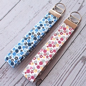 Pode incluir: Dois chaveiros florais. Um é azul com flores brancas e pretas, o outro é branco com flores rosa, amarelas e cinzentas. Cada um tem um anel de metal prateado e um fecho de metal dourado.