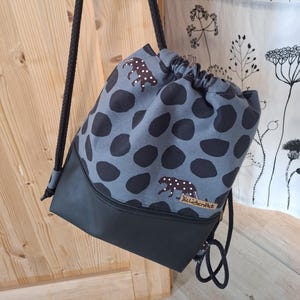 Puede incluir: Una mochila con cordón con una parte superior de tela gris con grandes lunares negros e ilustraciones de leopardo marrón. La parte inferior es negra, con correas de cuerda negra. La bolsa está colgada de una puerta de madera.