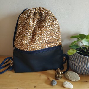 Könnte beinhalten: Ein Rucksack mit Kordelzug, mit einem Blumenmuster auf der oberen Hälfte und einem marineblauen Boden. Der Stoff ist beige mit kleinen weißen Blumen. Die Tasche hat einen marineblauen Reißverschluss und Kordelzüge. Der Text "DieLiebenäht" ist sichtbar.
