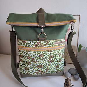 Könnte beinhalten: Grüne Canvas-Umhängetasche mit einer floral bedruckten Tasche und einem Rollverschluss. Die Tasche hat einen gelben Reißverschluss und einen verstellbaren Schultergurt. Ein Metallring und ein Verschluss sichern die obere Klappe.