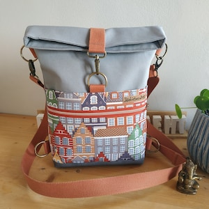 May include: Gray crossbody bag with a flap closure and a colorful cityscape pattern on the front pocket. The bag features a rust-colored strap and accents, with metal hardware. The bag has a zipper pocket and a roll-top design.