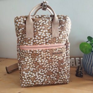 Könnte beinhalten: Ein brauner Rucksack mit Blumenmuster und weißen und braunen Blüten. Der Rucksack hat einen hellrosa Reißverschluss und beige Träger und Griffe. Der Text "Ateliernih" ist sichtbar.