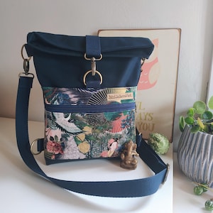 Könnte beinhalten: Eine marineblaue Umhängetasche mit Klappenverschluss und verstellbarem Riemen. Die Vorderseite hat eine Reißverschlusstasche mit einem farbenfrohen Blumen- und Vogelmuster. Die Tasche hat goldfarbene Hardware und einen kleinen dekorativen Anhänger.