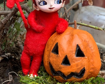 May include: A red felt devil doll with a heart-shaped tail stands next to a carved orange pumpkin with a jack-o'-lantern face. The doll and pumpkin are on a brown wooden base with green moss and white flowers.