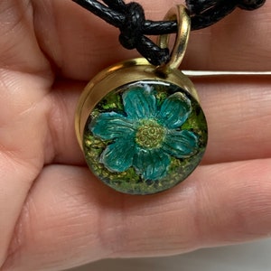 Könnte beinhalten: Eine goldene Halskette mit einem schwarzen Band. Der Anhänger zeigt eine blaue Blume, die in klarem Harz mit grünem und goldenem Hintergrund gefasst ist.