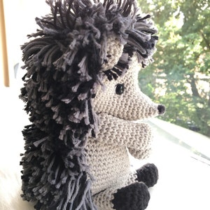 May include: A crocheted hedgehog plush toy with a gray and black body and black eyes. The hedgehog is sitting on a white surface.