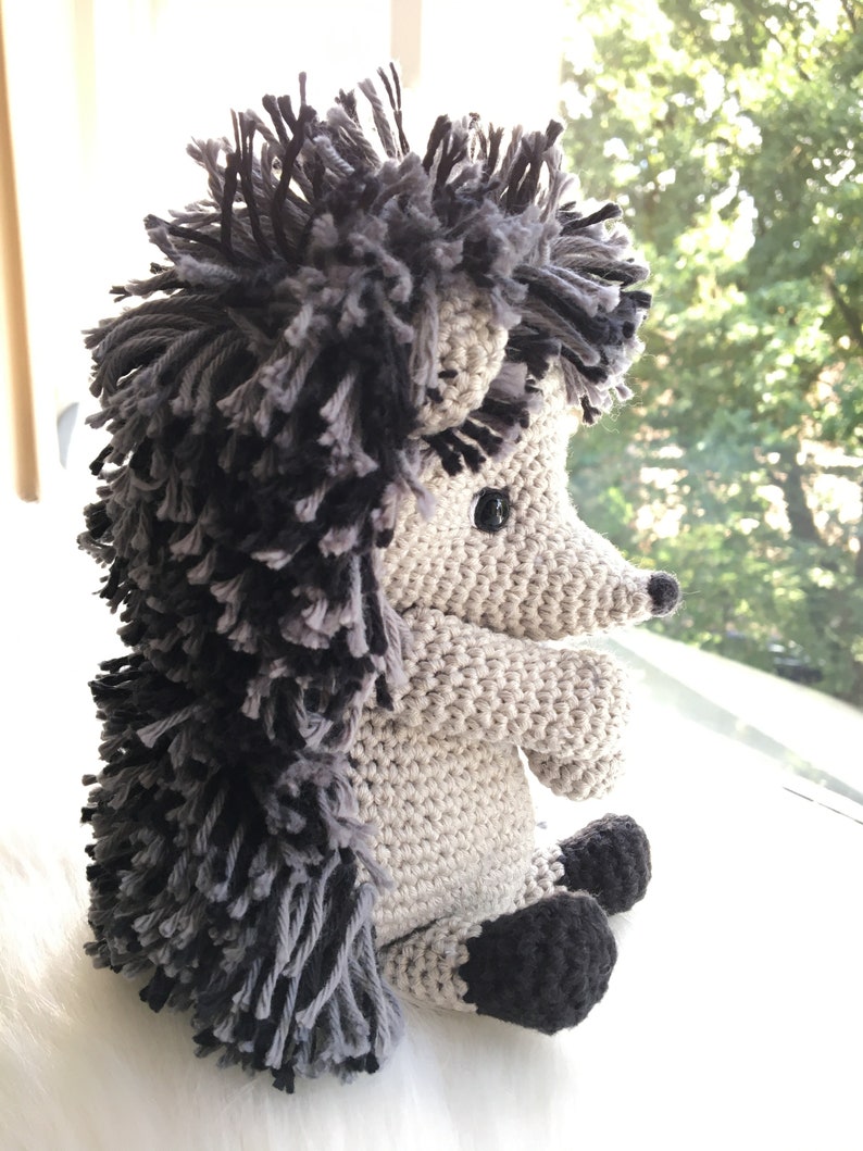 May include: A crocheted hedgehog plush toy with a gray and black body and black eyes. The hedgehog is sitting on a white surface.
