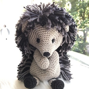 May include: A crocheted gray and black hedgehog plush toy with black eyes and a black nose. The hedgehog is sitting on a white fluffy surface.