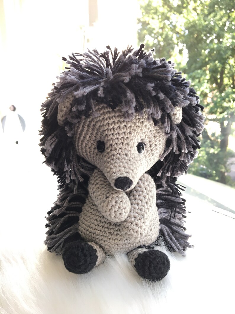 May include: A crocheted gray and black hedgehog plush toy with black eyes and a black nose. The hedgehog is sitting on a white fluffy surface.