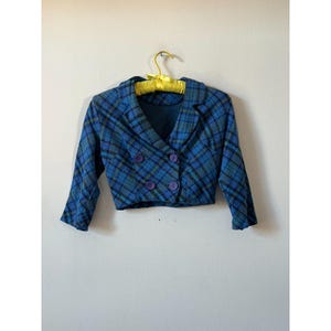 May include: A cropped blue plaid blazer with a double-breasted design. The blazer features a classic collar, three-quarter sleeves, and purple buttons. The jacket is hanging on a yellow hanger against a white wall.