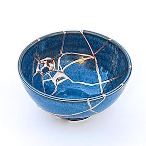 May include: A blue ceramic bowl with visible gold-colored kintsugi repair lines. The bowl has a rounded shape and a glossy finish. The kintsugi technique highlights the cracks with gold, creating a unique decorative piece.