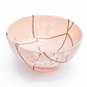 May include: A pink ceramic bowl with a floral design and gold-colored kintsugi repair lines. The bowl features a footed base and a delicate pattern of white flowers and stems. The kintsugi lines highlight the bowl's repaired cracks.