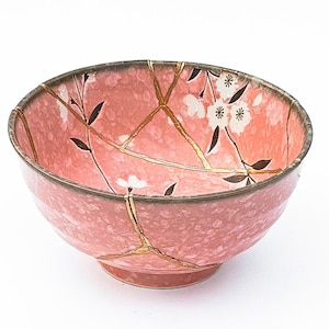 May include: A pink ceramic bowl with a floral design and gold accents. The bowl is cracked and repaired with gold kintsugi.