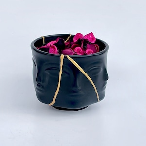 May include: Black ceramic pot with gold accents and a design of three faces. The pot is filled with pink potpourri.