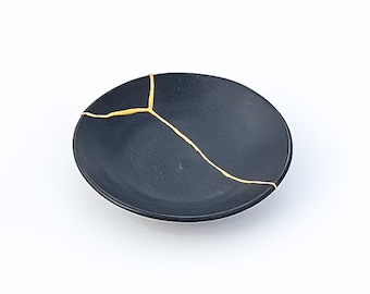 May include: A small, black ceramic dish with a crackled finish. The crack is filled with gold paint, creating a unique and decorative design.