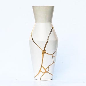 May include: A white vase with a tapered neck and a wider body, featuring a series of thin, raised gold lines that create a web-like pattern across the surface, resembling the kintsugi technique of repairing broken pottery with gold.