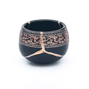 May include: A black ceramic bowl with a copper-colored decorative pattern around the top and copper-colored accents. The bowl has a unique shape and design, showcasing a blend of traditional and modern aesthetics.