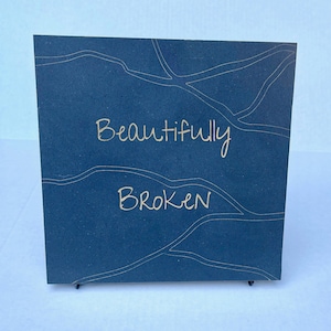 May include: A dark blue square sign with gold lettering that reads "Beautifully Broken" on a white background.