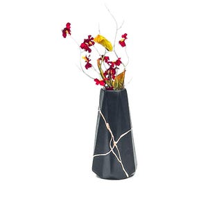 May include: A black, geometric vase with gold-colored kintsugi-style repairs, holding a floral arrangement. The arrangement features red and yellow flowers with thin, white branches. The vase has a unique, angular shape.