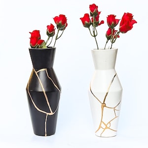 May include: Two tapered vases, one matte black and one matte white, each featuring gold-coloured lines resembling kintsugi repair patterns. Both vases contain artificial red rose stems with green leaves extending from the narrow openings.