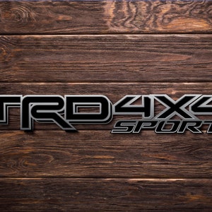May include: A black and silver 3D emblem with the text "TRD 4X4 SPORT" on a brown wooden background.